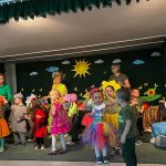 Grupa dzieci w wiosennych strojach z opiekunkami na scence teatralnej.