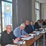 Grupa dorosłych osób w eleganckich ubraniach na sali konferencyjnej. Na stole znajdują się dokumenty i butelki wody.