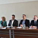 Trójka dorosłych męzczyn w graniturach i elegancko ubrana kobieta siedzacy za podłużnym stołem.