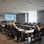 Grupa dorosłych osób w eleganckich ubraniach na sali konferencyjnej. Na stole znajdują się dokumenty i butelki wody.