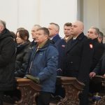 Grupa osób rożnej płci i wieku stojąca za drewnianymi ławkami kościelnymi .