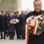 Mężczyzna elegancko ubrany trzymający bukiet kwiatów. W tle grupa ludzi wśród których stoi trzech mężczyzn w mundurach, jeden z nich trzyma bukiet kwiatów.