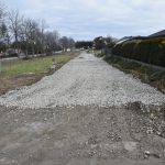 Utwardzona droga bez powierzchni asfaltowej obok domów mieszkalnych i ogrodzenia z żywopłotu.