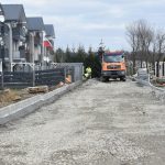 Droga utwardzona przy której stoi duży samochód i paru pracujacych mężczyn. Obok znajdują się domy mieszkalne.