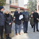 Grupa dorosłych i młodzieży stojacych przed gmachem urzędu. Starszy mężczyzna przemawia za pomocą mikrofonu.