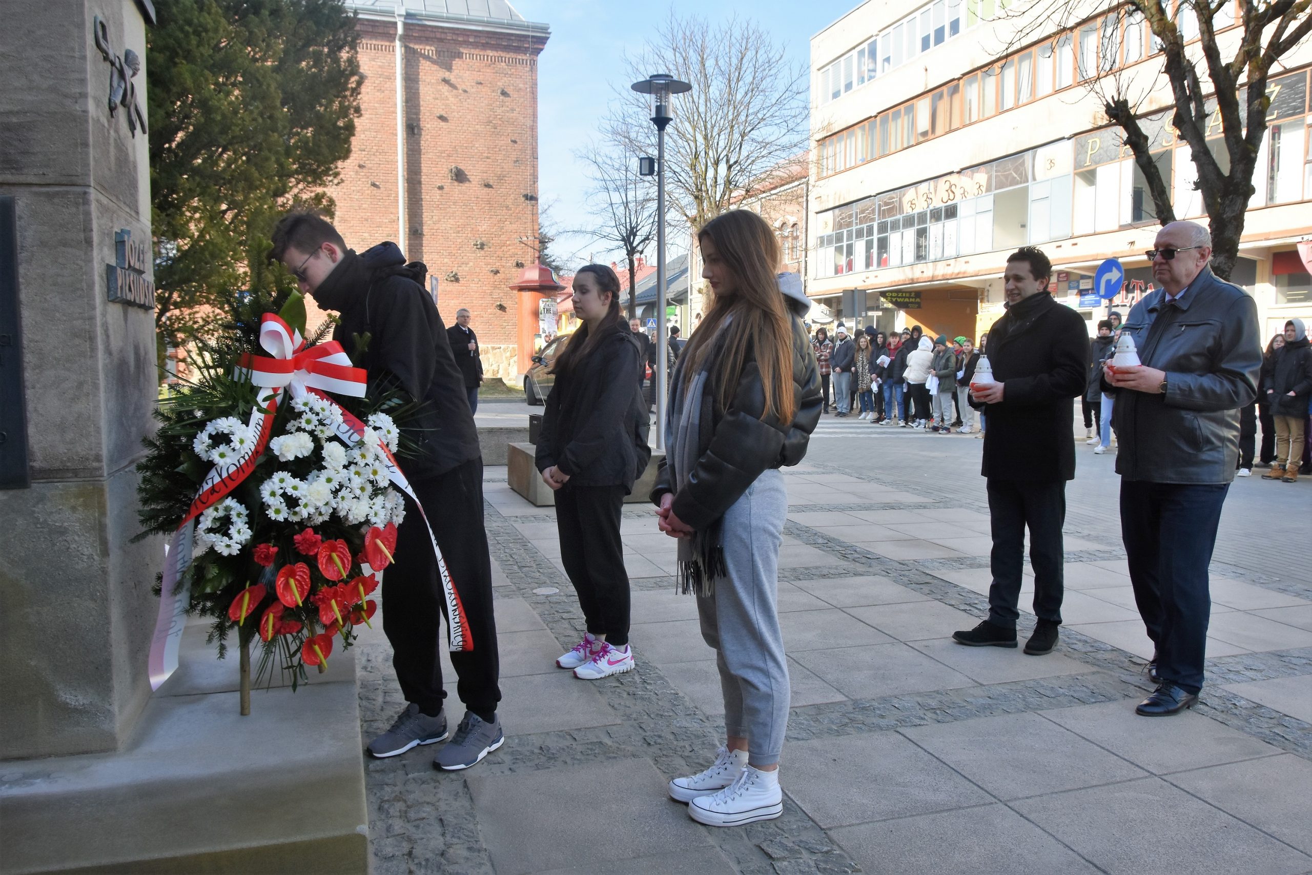 Grupa młodzieży i dwójka mężczyżn skłądajacych wieniec i znicze przed popiersiem Piłsudskiego.