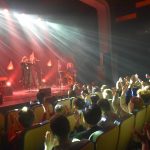 Młoda wokalistka i dwóch dorosłych mężczyzn w czasie występu na scenie. Ogląda ich publiczność siedząca na krzesłach w sali kinowej.