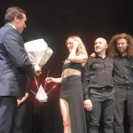 Mężczyzna w graniturze przekazuje bukiet kwiatów młodej wokalistce. Obok dóch mężczyzn w ciemnych strojach.