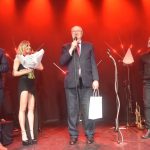 Trzech dorosłych mężczyn i młoda kobieta na scenie z kwiatami.