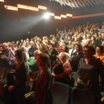 Duża grupa róznej płci i wieku stojaca na sali kinowej.