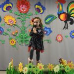 Dziewczynka na podeście przemawia za pomocą mikrofonu. Z tyłu widać zasłonę z ludowymi zdobieniami.