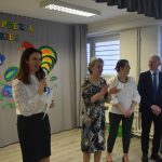 Trzy kobiety i elegancko ubrany starszy mężczyzna stojący na sali. Jedna z kobiet przemawia za pomocą mikrofonu.