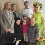 Starszy mężczyzna w białej koszuli pozuje do zdjecia z grupą dzieci i kilkoma kobietami. Jedna z nich ubrana jest w strój reprezentujacy wiosnę.