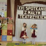 Dwójka dziecząt trzymajace kartonowe wizerunki ptaków. Obok stoi kobieta czytająca książkę.