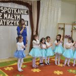 Grupa dziewcząt podzas wystepu tetralnego. Przy nich stoi kobieta przemaiwajaca za pomocą mikrofonu.