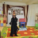 Dorosła kobieta w stroju czarownicy. Obok znajduje sie kartonowy domek.