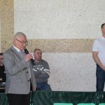 Starszy mężczyzna w szarym garniturze przemawiajacy na sali gimanstycznej. W tle widać kilku słuchajacych go mężczyzn.