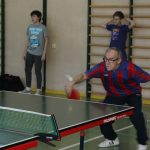 Starszy męzczyzna grajacy w tenis stołowy. Ogląda go dwoch młodych chłopaków. Wszyscy znajdują się na sali gimnastycznej.