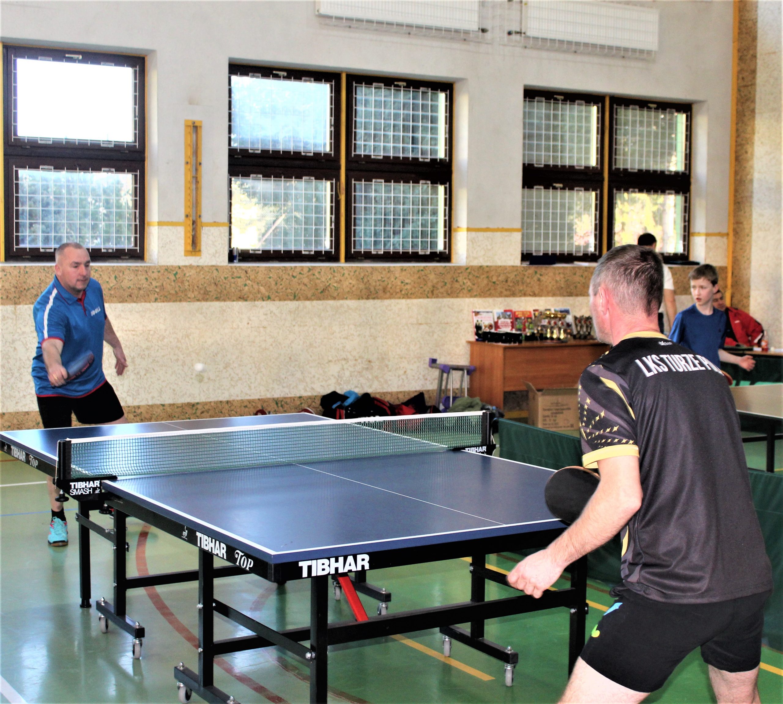 Dwóch mężczyzn na sali gimanstycznej grajacy w tenis stołowy.