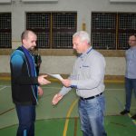 Mężczyzna w jasnej koszuli i jeansach przekazuje dyplom lauratowi konkursu.