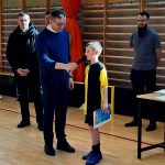 Grupa dorosłych mężczyzn z młodym zawodnikiem trzymajacym statuetkę oraz dyplom. Chłopak przemawia do mikrofonu.