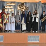 Grupa młodziezy na scenie w sali w czasie występu jasełkowego. Dwóch chłopców ubranych w garnitury trzyma instrumenty muzyczne.