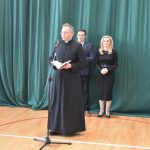 Ksiądz przemawiający przez mikrofon. W tle stoi kobieta i mężczyzna