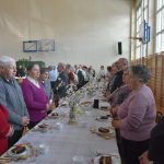 Grupa starszych osób róznej płci i wieku stojacy przy długim stole na sali gimanstycznej.