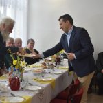 Ludzie siedzący przy stole z jedzeniem. Mężczyzna w garniturze częstuje jajkiem