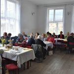 Sala z dużymi oknami w której siedzą ludzie przy zastawionych stołach.