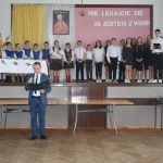 Grupa dzieci obranych w odswietne stoje na scenie i męzczyzna przemaiwjacy za pomocą mikrofonu.