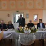 Grupa meżczyzn i kobiet zasiadajaca na sali przy zastawionych stołach. Jeden z mężczyzn przemawia za pomocą mikrofonu.