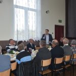 Grupa meżczyzn i kobiet zasiadajaca na sali przy zastawionych stołach. Jeden z mężczyzn przemawia za pomocą mikrofonu.