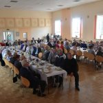 Duża grupa osób róznej płci i wieku zasiadajaca przy stołach na sali.