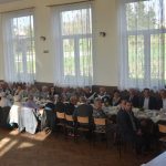 Duża grupa osób róznej płci i wieku zasiadajaca przy stołach na sali.