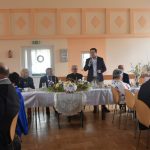 Grupa meżczyzn i kobiet zasiadajaca na sali przy zastawionych stołach. Jeden z mężczyzn przemawia za pomocą mikrofonu.