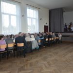 Grupa meżczyzn i kobiet zasiadajaca na sali przy zastawionych stołach. Jeden z mężczyzn przemawia za pomocą mikrofonu.