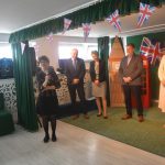 Grupa dorosłych męczyzn i kobiet stojacych na sali. Jedna z kobiet przemawia za pomocą mikrofonu.