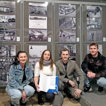 Dwie kobiety i dwóch mężczyzn prezentujący dyplom na tle ścianki ze zdjęć.