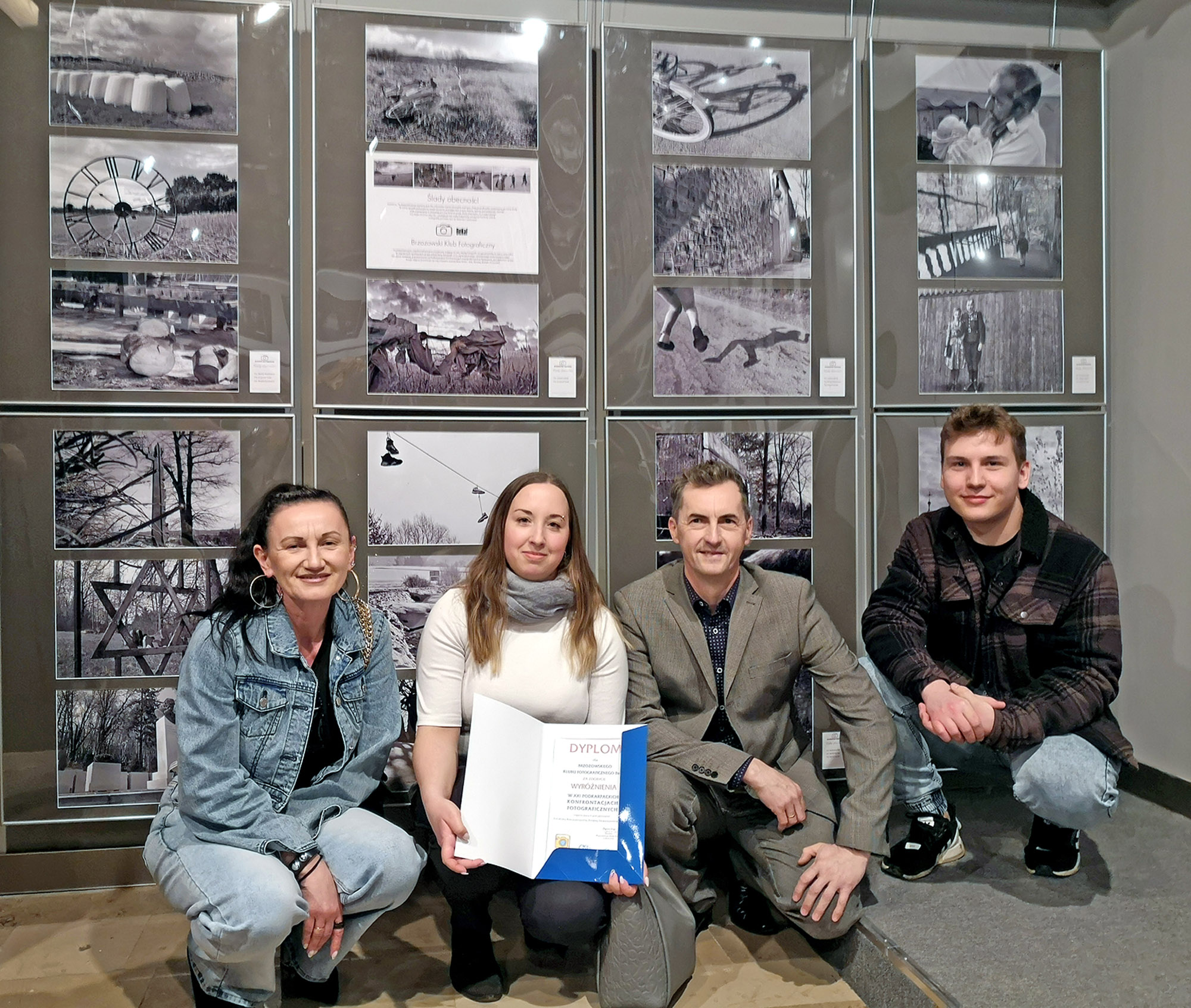 Dwie kobiety i dwóch mężczyzn prezentujący dyplom na tle ścianki ze zdjęć.