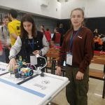 Grupa młodzierzy na rozległej sali. Na stole na pierwszym planie znajduje się mały robot.