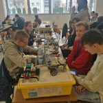 Grupa młodzieży siedzacej przy stoliku, na którym znajdują się roboty i cześci elektorniczne.