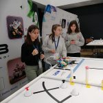 trójka dziewcząt przy stole na którym znajduje się niewielki robot.