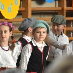 Grupa dzieci w odświetnych strojach na sali gimanstycznej w czasie przedstawienia wielkanocnego.