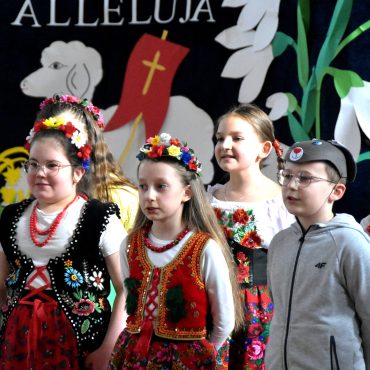 Grupa dzieci w odświetnych strojach na sali gimanstycznej w czasie przedstawienia wielkanocnego.