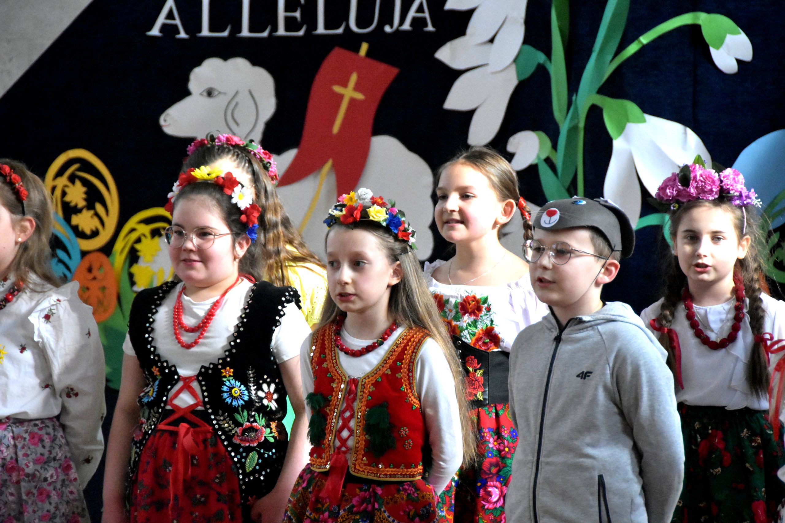 Grupa dzieci w odświetnych strojach na sali gimanstycznej w czasie przedstawienia wielkanocnego.