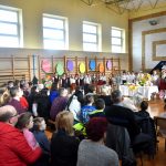 Grupa dzieci w odświetnych strojach na sali gimanstycznej w czasie przedstawienia wielkanocnego. Ogląda ich widowania złożona z osób rożnej płci i wieku.