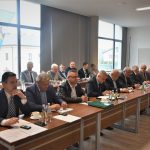 Grupa dorosłych osób w oficjalnych strojach siedzacych na sali obrad przy biurkach. Z tyłu widać okna z widokiem na miasto.