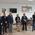 Grupa 5 dorosłych meżczyzn w graniturach i elegancko ubrana starsza kobieta na sali konferencyjnej. Jeden z mężczyzn przemawia za pomocą mikrofonu.