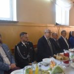 Grupa elegancko ubranych osób zasiada przy bogato zastawionym stole na sali.